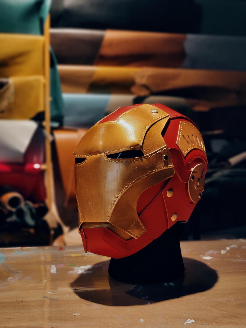 May include: A handcrafted red and gold leather helmet, reminiscent of a superhero's mask. The helmet features a gold faceplate, red accents, and decorative gold rivets. The design is detailed and appears to be a cosplay item.