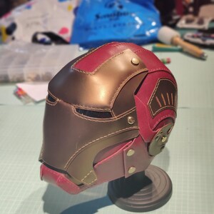 May include: A handmade leather helmet in shades of burgundy and brown, with brass-colored rivets and stitching. The helmet is displayed on a black stand, showcasing its intricate design and craftsmanship. The overall theme is cosplay or fantasy.