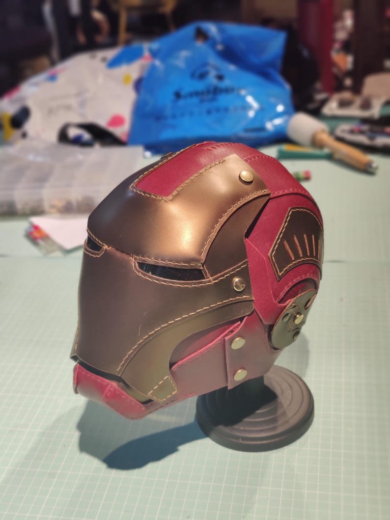 May include: A handmade leather helmet in shades of burgundy and brown, with brass-colored rivets and stitching. The helmet is displayed on a black stand, showcasing its intricate design and craftsmanship. The overall theme is cosplay or fantasy.