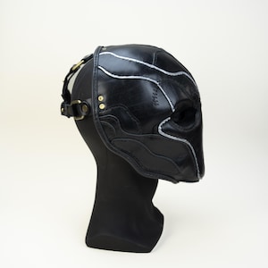 May include: Black leather mask with silver accent stitching and brass hardware. The mask is designed with a sculpted shape and features eye openings. It is displayed on a black mannequin head against a white background.