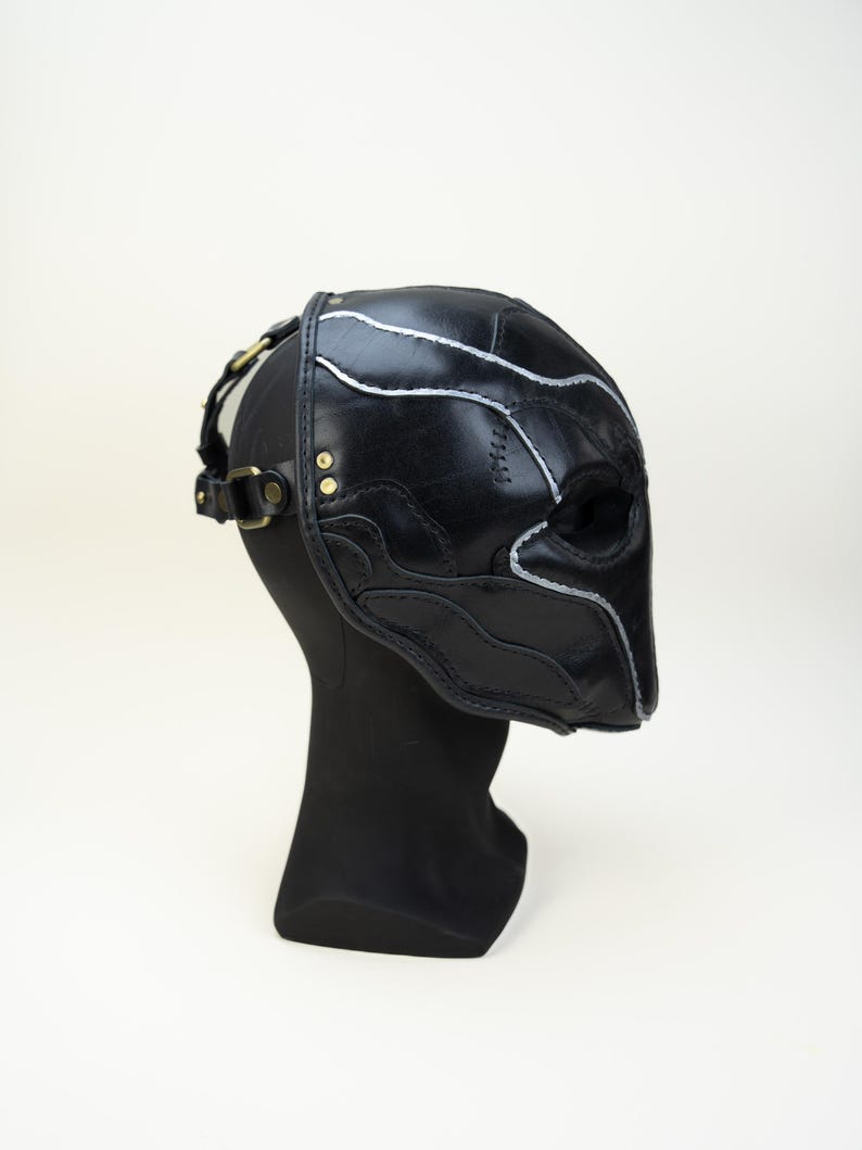 May include: Black leather mask with silver accent stitching and brass hardware. The mask is designed with a sculpted shape and features eye openings. It is displayed on a black mannequin head against a white background.