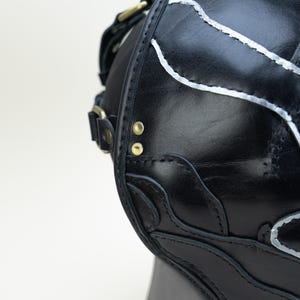 May include: Black leather satchel with silver accent stitching and brass hardware. The bag features decorative cutouts and a shoulder strap. The bag is shown against a white background.