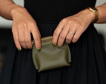 May include: A small, olive green leather coin purse with green stitching. The purse is held in the hands of a person wearing a black dress.