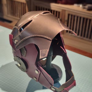 May include: A handmade leather helmet in shades of brown and purple, with visible stitching and metal accents. The helmet features a visor and ear coverings, suggesting a protective or decorative function. The design is intricate and detailed.