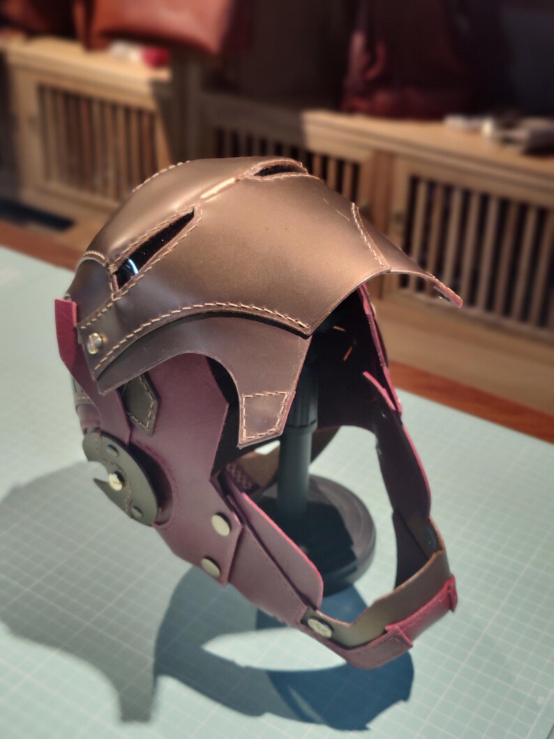 May include: A handmade leather helmet in shades of brown and purple, with visible stitching and metal accents. The helmet features a visor and ear coverings, suggesting a protective or decorative function. The design is intricate and detailed.