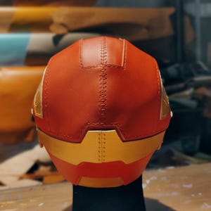 May include: A red and gold helmet with a stitched seam down the center. The helmet has gold accents and is displayed on a black stand. The back of the helmet is visible, showcasing the craftsmanship and design.