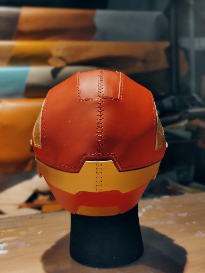 May include: A red and gold helmet with a stitched seam down the center. The helmet has gold accents and is displayed on a black stand. The back of the helmet is visible, showcasing the craftsmanship and design.