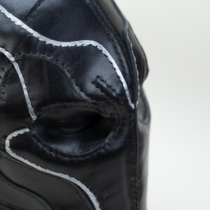 May include: Close-up of a black leather mask with silver stitching. The mask has an eye opening and is designed with curved lines and panels. The stitching follows the contours of the mask, creating a detailed and textured appearance.