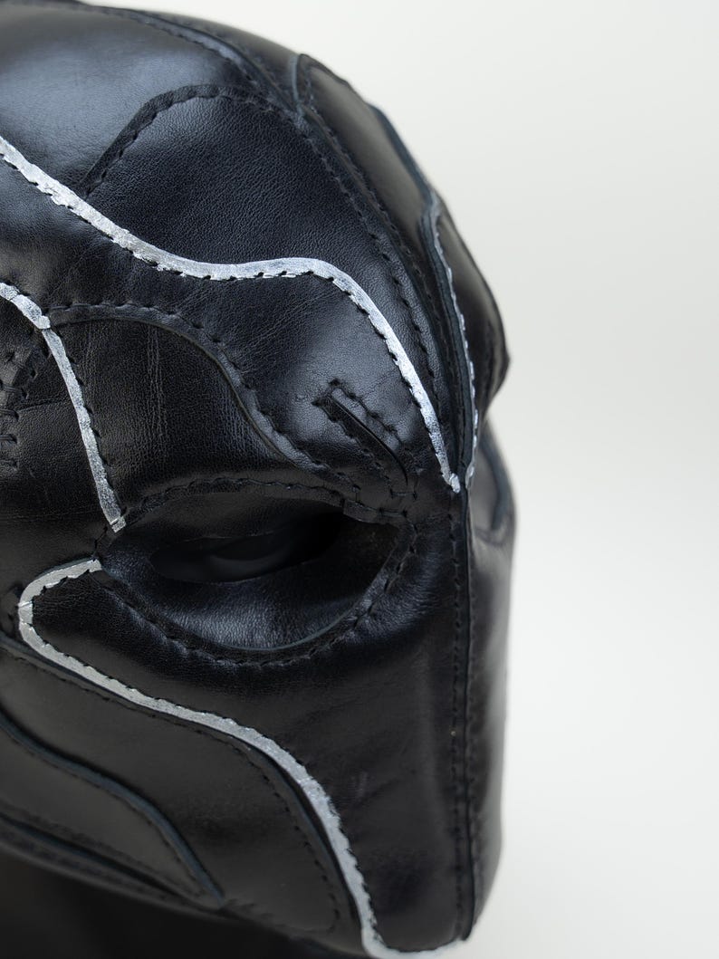 May include: Close-up of a black leather mask with silver stitching. The mask has an eye opening and is designed with curved lines and panels. The stitching follows the contours of the mask, creating a detailed and textured appearance.