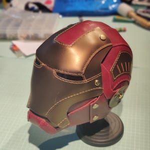 May include: A handcrafted, leather helmet in shades of brown and burgundy, featuring intricate stitching and metallic accents. The helmet is displayed on a black stand, showcasing its detailed design and craftsmanship.