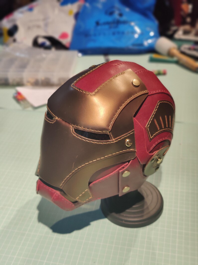 May include: A handcrafted, leather helmet in shades of brown and burgundy, featuring intricate stitching and metallic accents. The helmet is displayed on a black stand, showcasing its detailed design and craftsmanship.