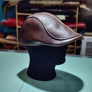 Puede incluir: Gorra plana de cuero marrón oscuro con visera curva, exhibida sobre una cabeza de maniquí negra. La gorra presenta costuras visibles y una superficie texturizada. El fondo muestra un taller con materiales de cuero.