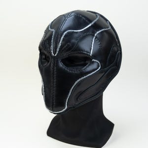 May include: Black leather mask with silver accents and stitching. The mask has eye openings and a contoured design. It is displayed on a black mannequin head against a white background. The mask's design features curved lines and shapes.