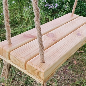 Può includere: Un'altalena in legno con tre assi, collegata da una spessa corda naturale. L'altalena è realizzata in legno di colore chiaro ed è sospesa su uno sfondo di erba verde e fogliame. L'altalena è progettata per uso esterno.