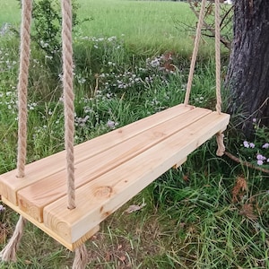 Può includere: Un sedile a dondolo in legno appeso a corde spesse, sospeso da un ramo d'albero. Il sedile è fatto di assi di legno naturale e ha un aspetto rustico e fatto a mano.