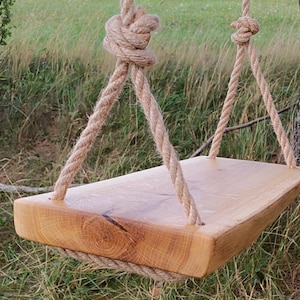Puede incluir: Un columpio de madera con acabado natural cuelga de gruesas cuerdas anudadas. El asiento del columpio es una tabla rectangular de madera clara, suspendida en un entorno exterior cubierto de hierba cerca del tronco de un árbol.
