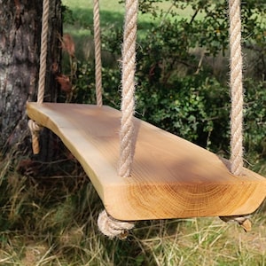 Könnte beinhalten: Eine Holzschaukel mit natürlicher Kante, aufgehängt an dicken, beigefarbenen Seilen. Der Schaukelsitz besteht aus hellem Holz, das die Maserung und Textur zeigt. Die Schaukel hängt an einem Baum in einer natürlichen Umgebung.