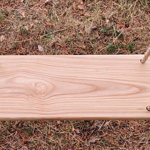 Könnte beinhalten: Ein hölzerner Schaukel Sitz mit natürlicher Holzmaserung, aufgehängt an dicken, beigefarbenen Seilen. Der rechteckige Sitz ist aus hellem Holz gefertigt und für den Außenbereich konzipiert. Die Schaukel befindet sich vor einem Hintergrund aus Gras und trockenem Laub.