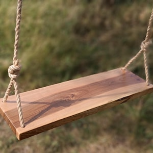 Könnte beinhalten: Eine Holzschaukel mit rechteckigem Sitz und Seilaufhängungen. Der Sitz besteht aus hellbraunem Holz mit einem natürlichen Maserungsmuster. Die Seile sind hellbeige und die Schaukel hängt vor einem verschwommenen grünen Hintergrund.