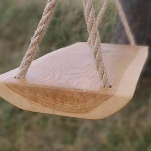 Könnte beinhalten: Eine Holzschaukel mit natürlicher Oberfläche, aufgehängt an einem dicken, geflochtenen Seil. Der Schaukelsitz hat eine geschwungene Kante und sichtbare Holzmaserung. Der Hintergrund ist unscharf, was auf eine Außenumgebung hindeutet.