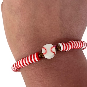 May include: A baseball-themed bracelet featuring red and white striped beads and a baseball charm. The charm is white with red stitching. The bracelet is worn on a wrist.