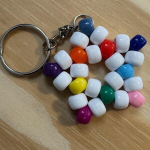 May include: A star-shaped keychain made of white and colorful beads. The beads are arranged in a star pattern and attached to a silver-colored keyring and chain. The beads are various colors, including blue, red, orange, and pink.