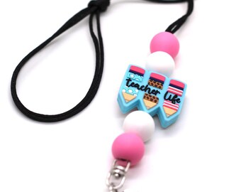 Puede incluir: Un cordón negro con un dije de cuentas rosa y blanco con tres lápices coloridos con el texto "teacher life". El cordón tiene un clip giratorio plateado y un anillo plateado.