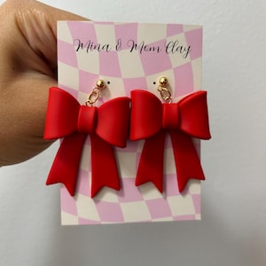 Puede incluir: Pendientes de lazo rojo con postes dorados y un detalle de cadena. Los pendientes se muestran sobre un fondo a cuadros rosa y blanco con el texto "Mina & Mom Clay".