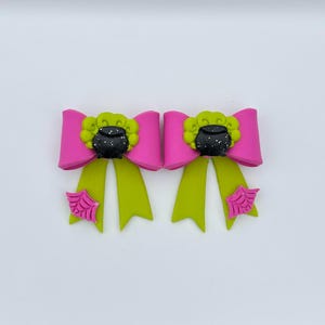 Pode incluir: Dois laços de cabelo com tema de Halloween. Cada laço apresenta um laço rosa brilhante com um centro e fitas verde limão. Um caldeirão preto com um redemoinho verde fica em cima do laço. Uma teia de aranha rosa é presa às fitas.
