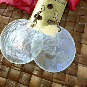 Può includere: Un paio di orecchini a conchiglia bianca iridescente con ganci dorati. Gli orecchini sono rotondi e hanno una texture naturale e organica.