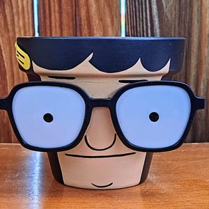 May include: A hand-painted ceramic planter shaped like a cartoon character with black hair, a brown nose, and black-rimmed glasses. The planter has a yellow stripe on the side.