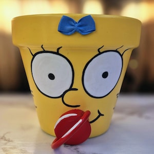 May include: Yellow painted flower pot with a cartoon face design. The pot features large white eyes with black outlines, a blue bow, and a red pacifier-shaped accent. The pot is designed to resemble a cartoon character.