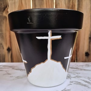 Peut inclure: Pot de fleur peint en noir avec des croix blanches et une base blanche. Les croix ont un contour beige. Le pot a un large bord et un corps légèrement conique. L'arrière-plan est une clôture en bois.