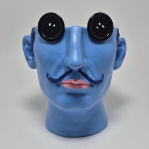 May include: A light blue head-shaped planter featuring two large black four-hole buttons as eyes, a dark blue sculpted mustache, and light pink lips. The object is hollow at the top, designed to hold items or plants within the head structure.