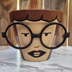 Puede incluir: Maceta de cerámica pintada a mano con diseño de rostro. La maceta presenta una cara marrón claro con gafas de montura negra, boca negra y cabello castaño. Diseñada para plantas y flores.