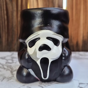 May include: A black ceramic mug featuring the iconic Ghostface mask from the Scream horror film series. The mask is white with black eye and mouth details, set against a glossy black background. The mug has a rounded base and a cylindrical shape.