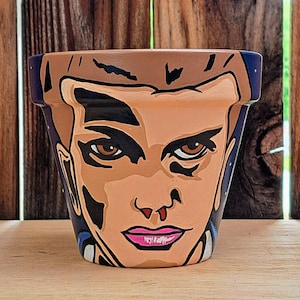 May include: A terracotta flower pot painted with a cartoon face. The face has brown hair, brown eyes, and a pink mouth. The pot is painted in a blue and black colour scheme.