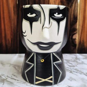 Könnte beinhalten: Eine Keramik-Tasse mit einem stilisierten Gesicht in Schwarz und Weiß bemalt. Das Gesicht hat schwarzes Augen-Make-up, einen schwarzen Mund und einen schwarzen Anzug mit goldenem Akzent. Die Tasse ist zylindrisch und hat eine glatte Oberfläche.