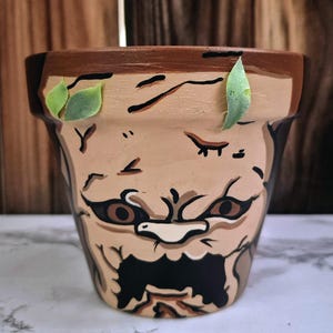May include: Hand-painted terracotta flower pot with a face design. The pot features a tan base with brown and black details, including eyes, a nose, and a mouth. Two green leaves are attached to the rim. The pot is designed for planting flowers or succulents.