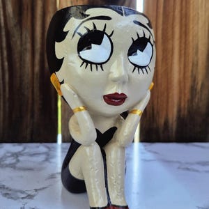 May include: A ceramic planter shaped like a seated cartoon woman with a black dress and red shoes. The planter has a cream-colored face with black hair, large eyes, and red lips. Gold accents are on the ears and arms.