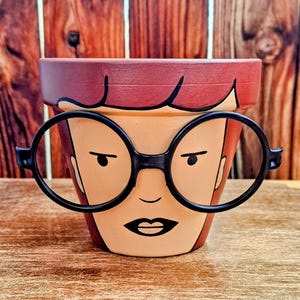 Pode incluir: Um vaso de flores de terracota pintado com um rosto de desenho animado usando óculos redondos pretos. O rosto tem cabelo ruivo, olhos castanhos e uma expressão séria.