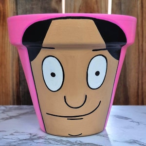 May include: A white ceramic mug with a black handle and the text "But First, Coffee" in a bold, black font. The mug is set against a blurred background, suggesting a cozy setting.