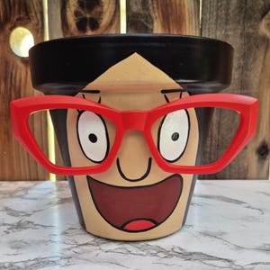 Pode incluir: Vaso de flores de cerâmica pintado à mão com um rosto de desenho animado com óculos vermelhos, boca sorrindo e cabelo preto. O vaso é bege com detalhes em preto e vermelho, perfeito para uso interno ou externo.