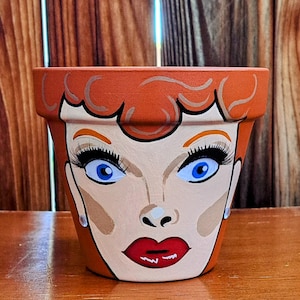 Könnte beinhalten: Ein Terrakotta-Blumentopf, bemalt mit einem Cartoon-Porträt einer Frau mit roten Haaren, blauen Augen und roten Lippen.