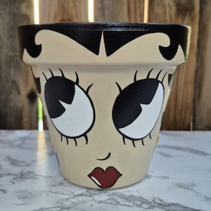 Puede incluir: Maceta de cerámica beige con una cara pintada a mano, cabello negro, ojos grandes y labios rojos. La maceta tiene un diseño de dibujos animados clásico, adecuado para uso en interiores o exteriores. Es un artículo decorativo.