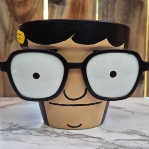 Peut inclure: Pot de fleurs en céramique peint à la main avec un visage de dessin animé. Il présente des lunettes à monture noire, un bandeau noir et une expression souriante. Le pot a un visage bronzé et un bord noir.