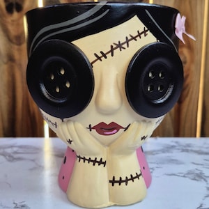 May include: A ceramic planter with a stylized face design. The face features large black button eyes, stitched details, and a pink bow. The color palette includes black, beige, pink, and red.