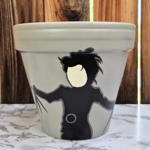 Peut inclure: Pot de fleur en céramique peint en gris clair, orné d'une silhouette noire d'un personnage aux mains de ciseaux. Le visage du personnage est de couleur beige clair. Le pot a un large bord et un corps légèrement conique.