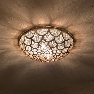 May include: A bronze-finished ceiling light fixture. The circular light has a detailed, patterned design. The light is on, casting a warm glow and intricate shadows on the ceiling. The light's diameter is approximately 40 cm.