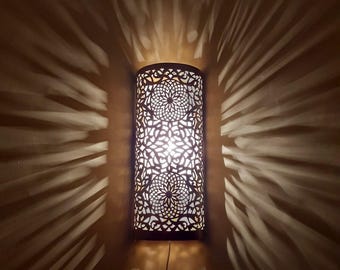 May include: A decorative wall sconce with a cylindrical shape. The sconce is made of dark metal with intricate, openwork patterns that cast a radial light pattern on the wall. The light source is visible through the cutouts.